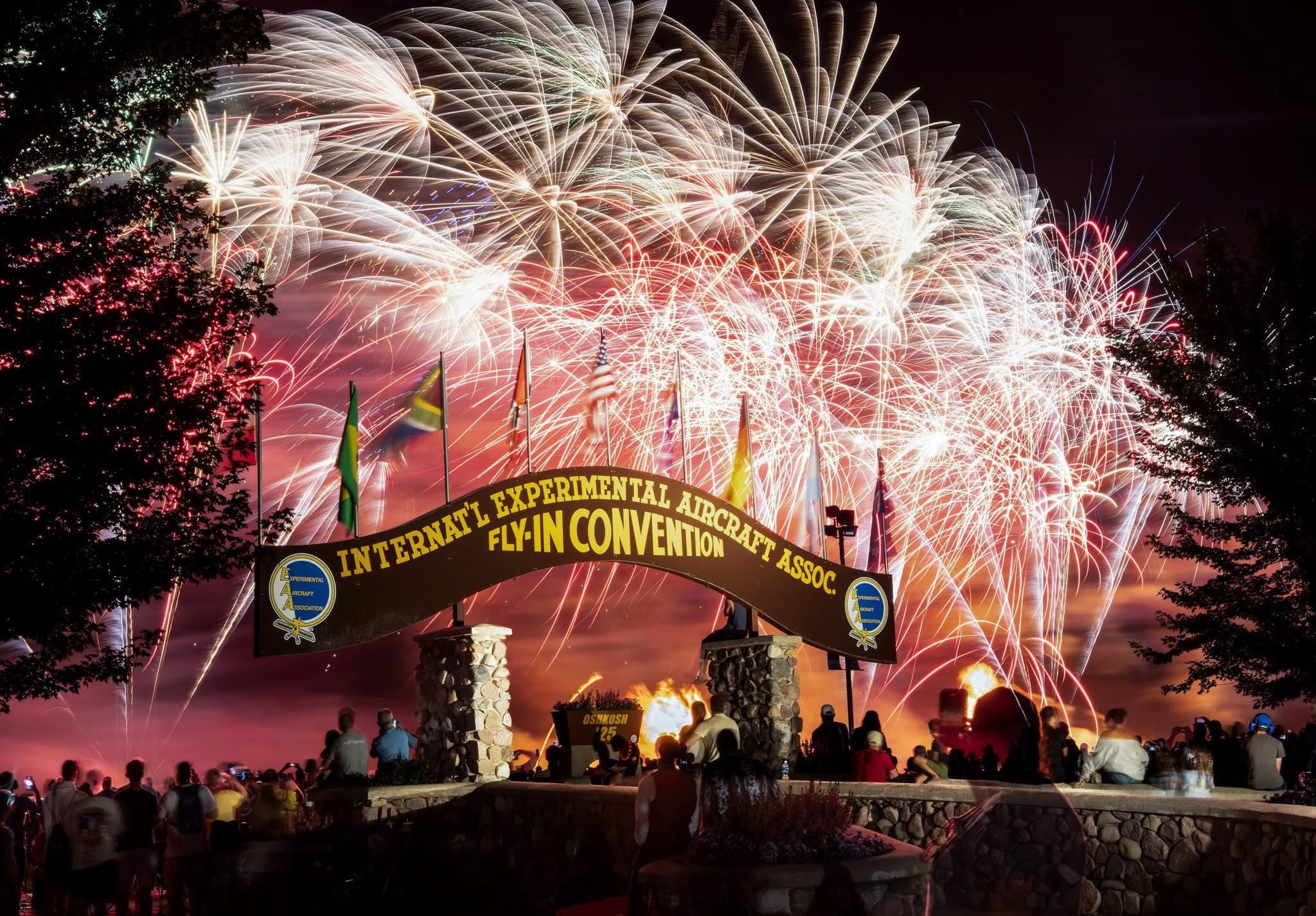 Fireworks bursting over the EAA AirVenture Oshkosh entrance arch at night with spectators gathered below (Shiny Jets DIY 3-day aircraft detailing training)