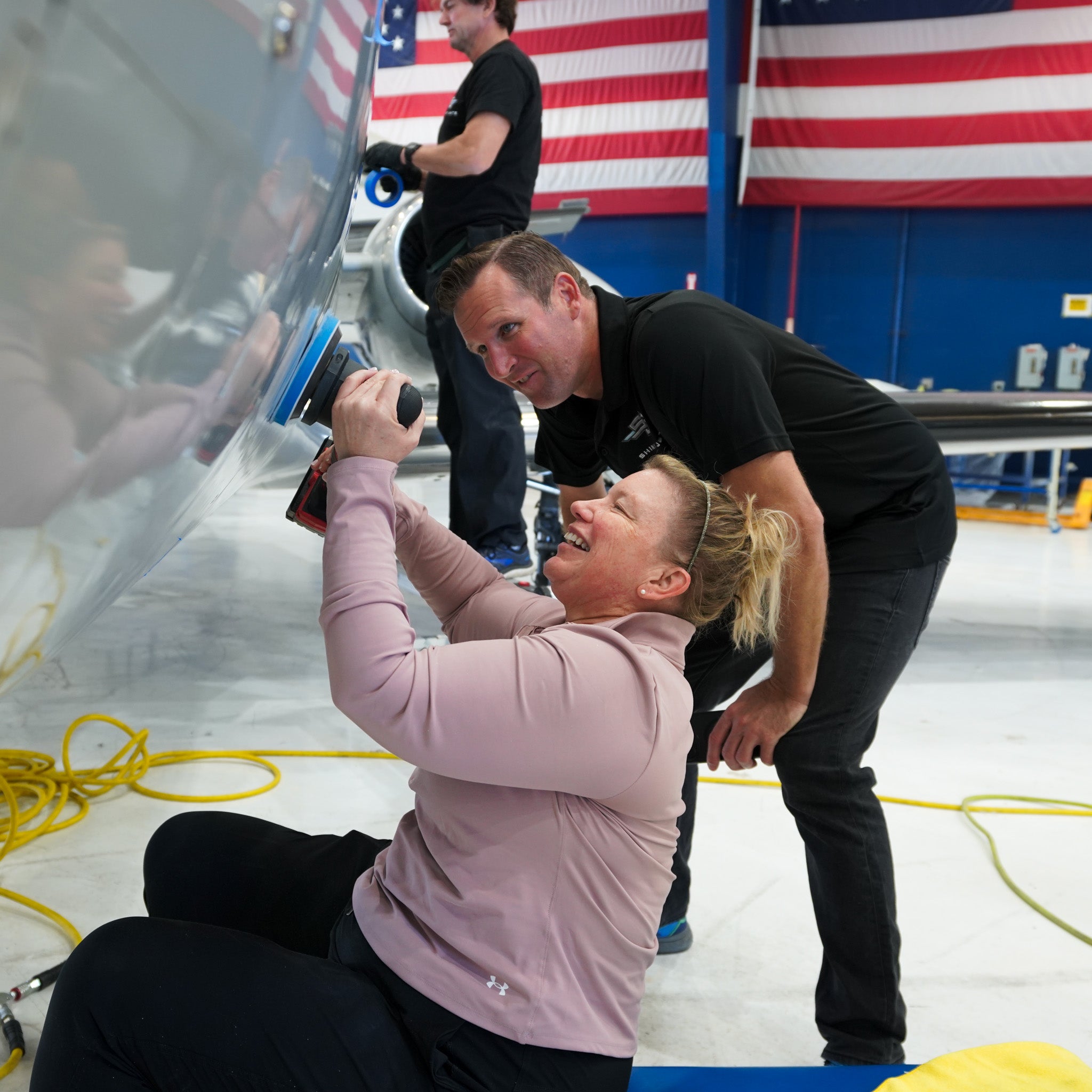 Shiny Jets DIY aircraft detailing training at EAA AirVenture Oshkosh: instructor coaching a student polishing an aircraft fuselage with a buffer inside a hangar (American flag in background).