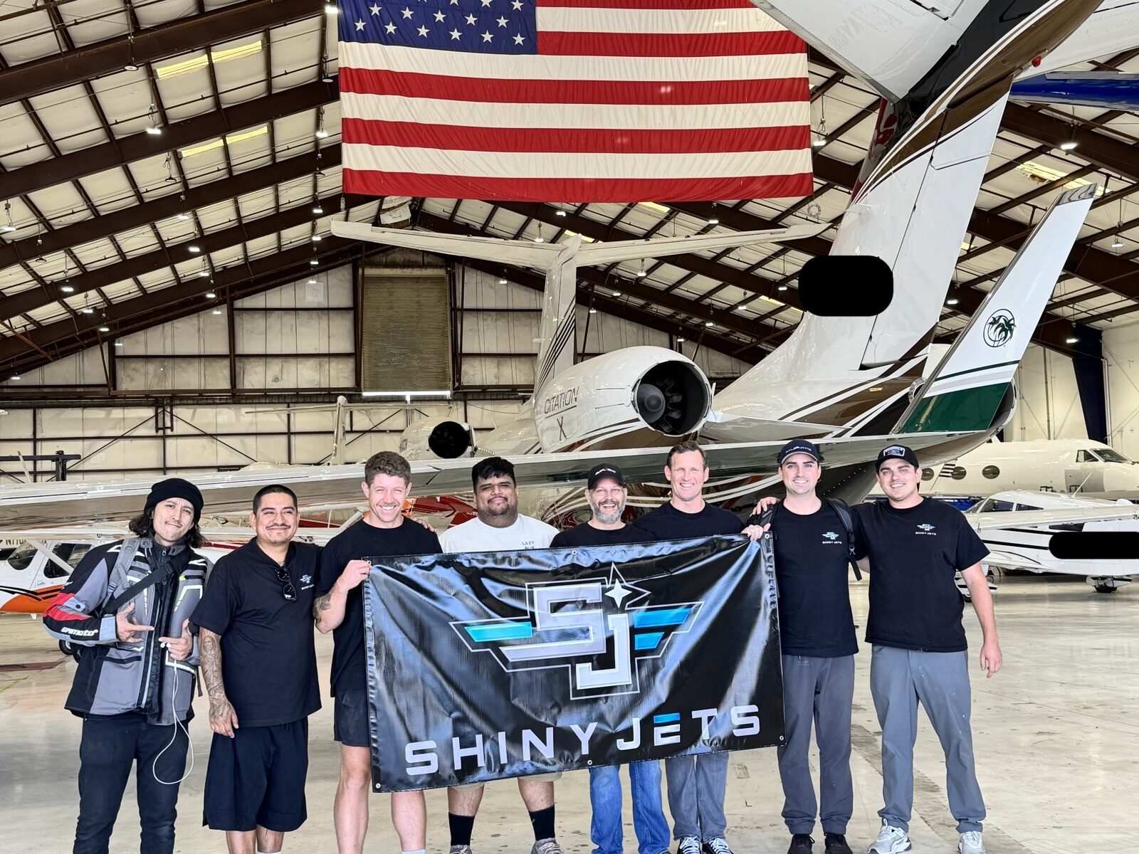 Shiny Jets team holding the SJ banner in a hangar full of business jets